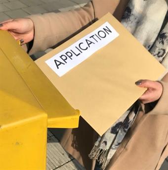 Eine Person in einem beigen Mantel ist dabei einen großen Briefumschlag, auf dem Application steht, in einen gelben Briefkasten zu werfen.