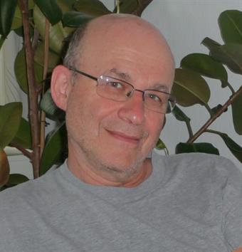 Ein älterer Herr lächelt in die Kamera. Er sitzt vor einer grünen Zimmerpflanze.