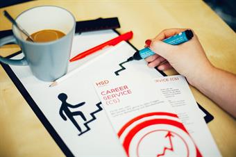 A filled coffe mug, set on a writing pad. Next to it two red and White leaflets of the career Service. A Hand is drawing with a black felt pen a stickman, who is climbing some steps.