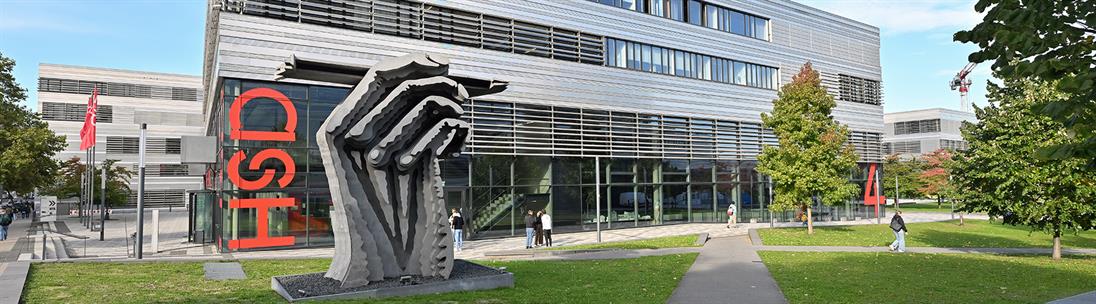 Außenansicht des Campus Derendorf der Hochschule Düsseldorf (HSD). Im Vordergrund steht eine markante, graue Metallskulptur auf einer Rasenfläche. Dahinter ist die moderne Glasfassade von Gebäude 4 zu sehen, an der vertikal der rote Schriftzug „HSD“ angebracht ist. Mehrere Personen laufen über die gepflasterten Wege des sonnigen Campus mit grünen Bäumen und Rasenflächen.