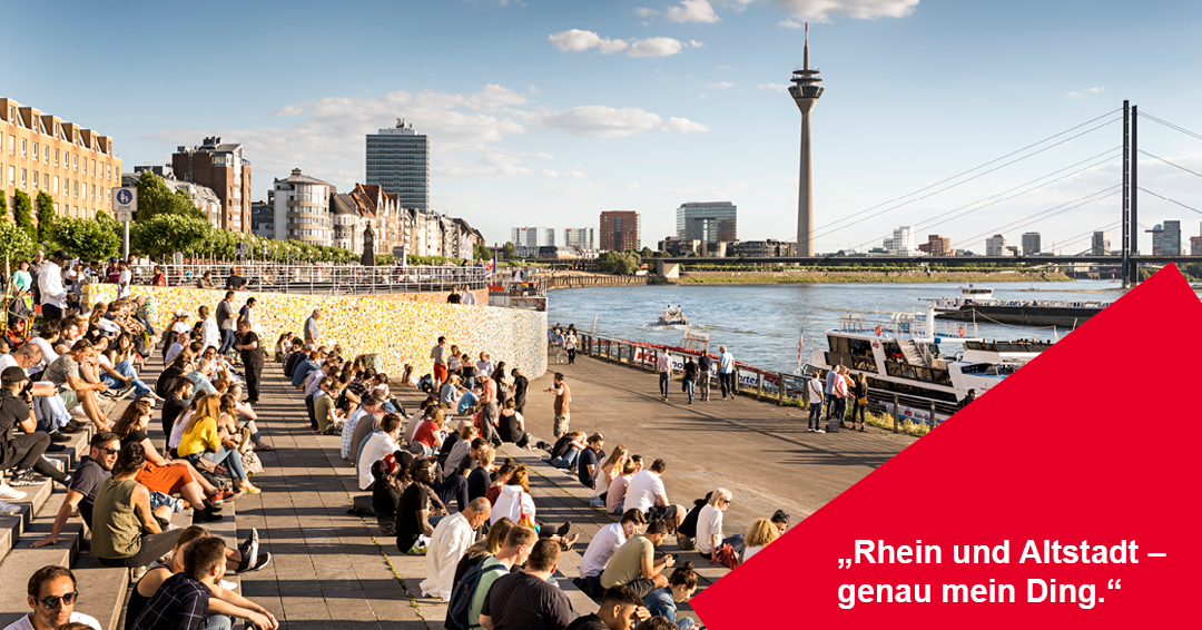 Eine Weitwinkel-Aufnahme der vollen Rheintreppe in Düsseldorf an einem sonnigen Nachmittag. Zahlreiche Menschen sitzen auf den breiten Steinstufen und genießen die Sonne.
