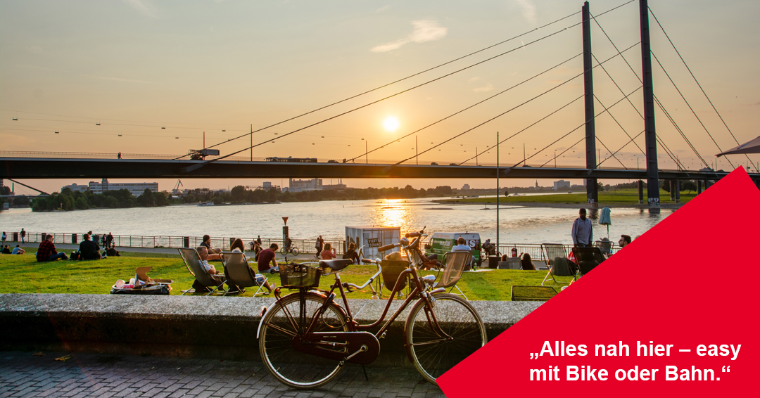 Ein stimmungsvolles Foto der Düsseldorfer Rheinwiesen bei Sonnenuntergang. Im Vordergrund steht ein rotes Vintage-Damenrad mit Korb an einer Mauer.
