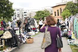Abgebildet ist ein Schotterweg, der umgeben ist mit Wiese auf der ein Flohmarkt stattfindet. Im Vordergrund eine Frau mit einem burgunderfarbendem Kleid, die verschwommen ist.
Rechts und links von ihr sind Kleidung, Möbel und weitere Menschen abgebildet.