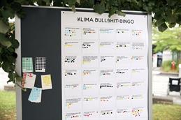 Abgebildet ist eine große Stoffpinnwand, welche draußen unter einem Baum aufgestellt ist. Die grünen Blätter des Baumes säumen den Bildrand oben. An der Pinnwand hängt ein großes Plakat mit der Aufschrift "Klima Bullshit-Bingo". Darunter viele gerahmte quadrate mit Sätzen zum Klimaschutz und bunten, runden, kleinen Aufklebern.