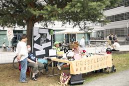 Abgebildet ist ein kleiner Stand draußen, an dem 4 ältere Frauen sitzen auf Stühlen, auf einer Wiese sitzen. Im Hintergrund ist ein Nadelbaum und die Metallfassade der Hochschule Düsseldorf zu sehen. Die Damen sind miteinander in ein Gespräch vertieft und auf dem Tisch des Standes stehen allerlei Dinge, die an einen Flohmarkt erinnern. Hinter den Frauen steht eine große Stoffpinnwand, an welche ein paar große Zettel geklebt sind.