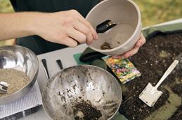 Abgebildet sind zwei Hände die einen Topf und eine kleine Schaufel halten, in der sich etwas Samengut befindet. Auf dem darunter stehenden Tisch stehen zwei weitere Schalen. Eine ist mit grauem Kies gefüllt, die andere wird als Mixschale genutzt, in der sich Erde, Kies und Samengut befindet. Rechts daneben ist eine Plastikplane, mit Erde.