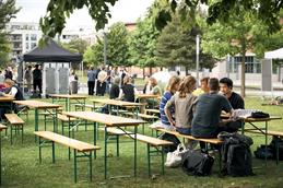 Abgebildet ist eine Wiese mit Bäumen und einem Wohngebiet im Hintergrund auf der Bierbänke mit -tischen stehen, auf denen viele junge Menschen sitzen, essen, trinken und miteinander sprechen. 