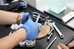 Abgebildet sind zwei Hände mit gummierten Arbeitshandschuhen. Die Person hat ein Cuttermesser in der rechten Hand und einen bunten Plastikkarabiner in der linken Hand. Die Klinge des Cuttermessers schneidet etwas Plastik vom Karabiner ab. Im Hintergrund ist verschwommen ein Tisch mit Werkzeug zu erkennen.