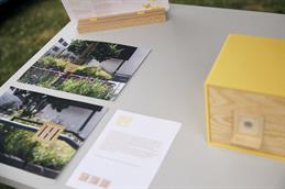 Abgebildet ist ein Tisch, von oben fotografiert, auf welchem zwei Bilder mit Blumen, Baum und einem gelben Würfel zu sehen sind. Im rechten Bildrandanschnitt ist der gelbe Würfel mit Holzfront und kleiner Plastikklappe vor einem runden Loch abgebildet.