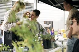 Abgebildet sind zwei junge Frauen, die vor einem Stand mit vielen Pflanzen stehen und zueinander gewandt sind. Die eine Frau streckt ihren Arm zu einer kleinen Box, in der Samengut in kleinen Tüten verpackt steht. Im rechten Bildrand ist ein sitzender, junger Mann mit Brille angeschnitten.
