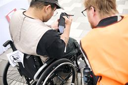 Abgebildet sind zwei Personen im Rollstuhl, die von oben über ihre Schultern fotografiert wurden. Der junge Mann links hat eine Kamera in der Hand und klickt Bilder durch, während der rechte Mann mit auf die Bilder schaut und eine orangene Warnweste trägt.