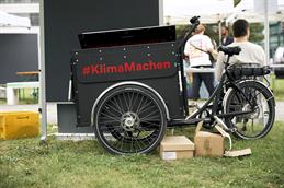 Abgebildet ist ein dreiradiges Fahrrad mit einer großen schwarzen Box vorn am Lenker, die den roten Schriftzug "#klimamachen" trägt. Das Fahrrad steht auf einer grünen Wiese und hinter diesem ist eine schwarze große Stoffpinnwand abgebildet. Unter ihm sind ein paar niedrige, kleine Kartons und im Hintergrund kann man verschwommen Rücken von Menschen erkennen.