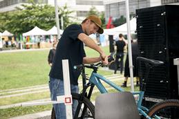 Abgebildet ist ein junger Mann mit brauner Cap, schwarzem weiten T-Shirt und einer Jeans, der konzentriert mit buntem Werkzeug vor einem Fahrradlenker steht und mit einem Inbusschlüssel am Lenker hantiert. Das Fahrrad ist ein Mountainbike mit türkisem Rahmen. Im Hintergrund zu sehen ist der Campus der Hochschule Düsseldorf und ihre grüne Wiese und die Metallfassade der Gebäude.