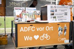 Abgebildet ist ein Stand des ADFC, welcher auf einem Fahrrad installiert wurde. Der Hintergrund der installierten Auflagefläche ist in einem hellen Orange gehalten. Darauf gestellt wurden viele Informationsflyer. Dahinter angeschnitten zu sehen sind zwei Personen, die zum ADFC gehören.