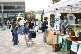 Abgebildet ist der gepflasterte Weg vom Campus der Hochschule Düsseldorf vor dem Gebäude 6. Es ist eine Essensbude und ein Stand zum Plastikrecycling im Fokus, welcher mit einem weißen Pavillon überdacht ist.