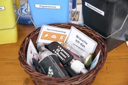 Abgebildet ist ein brauner Holztisch, auf dem ein geflochtenes Weidenkörbchen steht. In diesem Korb befinden sich alles Mögliche an Haushaktsmüll. So zum Bespiel Ein Schraubglas, eine LED-Birne, eine PET-Flasche, eine alte Fernbedienung, ein Korken und zwei Schilder, auf denen einmal "Sperrmüllsammlung" und "Brauche ich das wirklich", steht. Im Hintergrund stehen kleine Mülltonnen auf dem Tisch; eine gelbe, eine blaue mit der Aufschrift "Papiertonne" und eine schwarze mit Aufschrift "Restmülltonne".