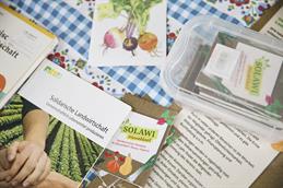Abgebildet ist eine blau-weiß karierte Tischdecke mit rot-orangenen Blumenelementen auf der einige Flyer zum Thema "Solidarische Landwirtschaft" steht.