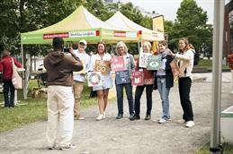 	
Abgebildet ist eine in Reihe stehender Personen, die zu einem Fotografen schauen und quadratische, bunte Banner festhalten. Sie stehen auf einem Schotterweg umgeben von Pavillons.
