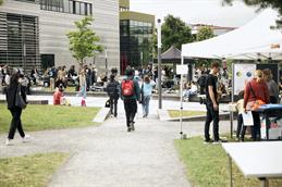 	
Abgebildet ist ein Schotterweg, der zu einem gepflasterten Bereich führt. Auf ebendiesem bewegen sich junge Menschen, die auf den im Hintergrund stattfindenden Flohmarkt laufen. Dort sitzen Menschen auf Decken auf der Wiese. Es ist sehr voll und gut besucht. Dahinter erkennt man die Gebäude der Hochschule Düsseldorf.