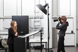 Eine schwarz gekleidete Fotografin fotografiert eine schwarz gekleidete Dozentin unter einem großen schwarzen Scheinwerfer vor einer weißen Wand