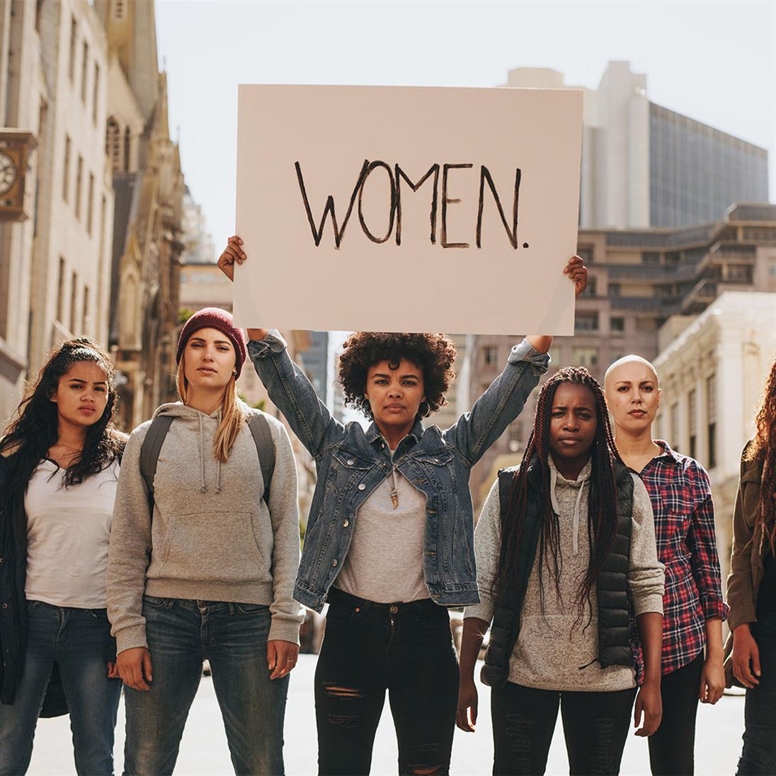 Fünf Frauen stehen in einer Reihe. Eine hält ein Schild hoch, auch welchem "Women" steht.