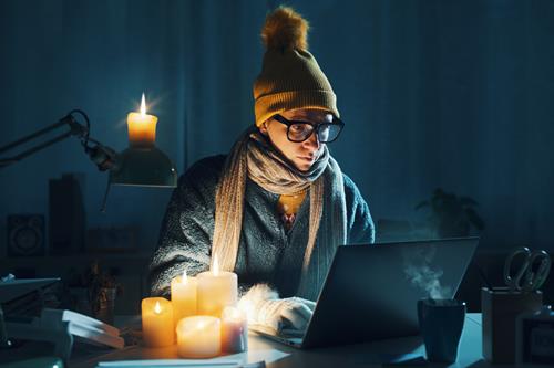 Eine Person mit Mütze und Handschuhen sitzt offenbar frierend vor ihrem Laptop. Rechts und links befinden sich Kerzen.