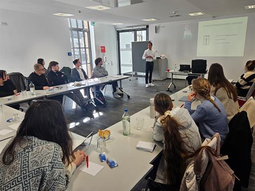 Ein heller Seminarraum mit Teilnehmern, die hufeisenförmig an Tischen sitzen. Der Trainer steht vorne an einer Leinwand mit der Überschrift „Unsere Körpersprache scannen“.