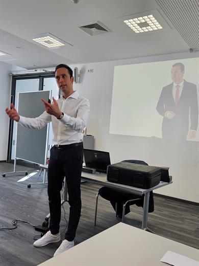 Ein männlicher Referent in weißem Hemd und dunkler Hose erklärt mit lebendiger Handgestik ein Thema. Im Hintergrund ist eine Beamer-Projektion eines Mannes im Anzug zu sehen.