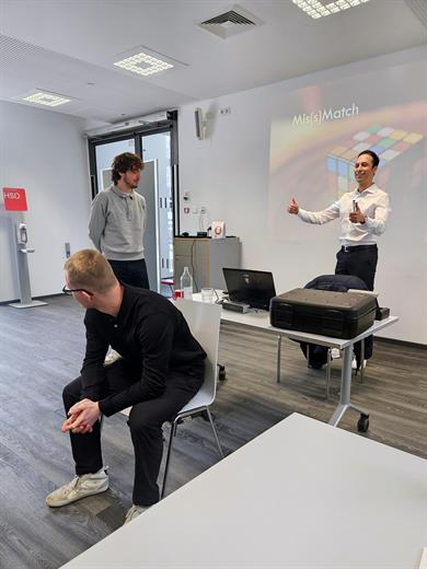 Ein lächelnder Coach steht vor einer Leinwand mit der Aufschrift „Mis(s)Match“ und einem Zauberwürfel-Motiv. Er zeigt zwei Daumen nach oben in Richtung der Teilnehmer.