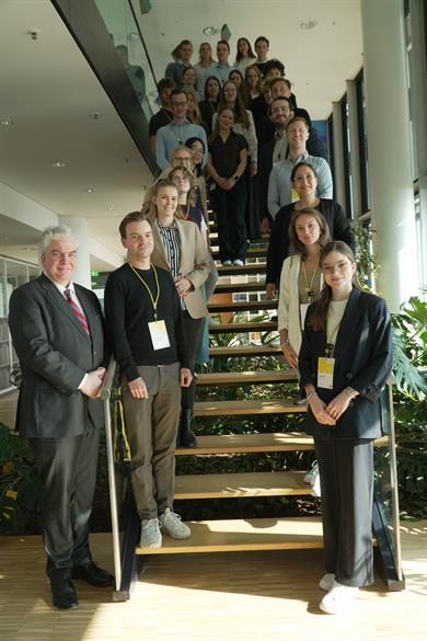 Das Bild zeigt eine Gruppe von Menschen, die in einem modernen Gebäude mit einer Treppe und viel natürlichem Licht posieren. Die Gruppe besteht aus mehreren Personen, die sowohl auf den Stufen als auch auf dem Podest der Treppe stehen. Die Menschen auf der Treppe sind in zwei Reihen angeordnet: eine Reihe steht auf den Stufen, während die andere Reihe auf dem Podest steht.