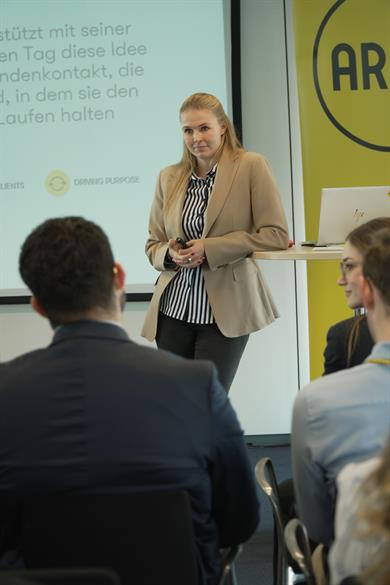 Eine Frau steht vor einer Gruppe von Menschen und hält einen Vortrag. Sie trägt einen beige-farbenen Blazer über einem schwarz-weißen gestreiften Hemd und hält ein Mikrofon in der Hand. Ihr Gesichtsausdruck ist konzentriert und professionell.
Im Hintergrund ist eine Projektionsfläche zu sehen. Neben der Projektionsfläche befindet sich ein gelbes Banner mit dem sichtbaren Logo und dem Wort "ARAG".
