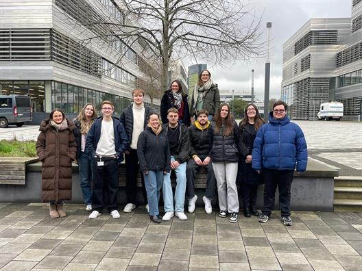 Eine Gruppe von 13 jungen Erwachsenen steht und sitzt in Winterkleidung auf einer niedrigen Betonbank im Freien auf einem modernen Campusgelände mit grau gepflastertem Platz, kahlem Baum und mehrstöckigen Gebäuden mit großen Glasfassaden im Hintergrund; der Himmel ist bewölkt.