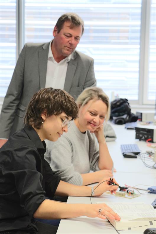 Mehrere Personen arbeiten gemeinsam an einem Tisch in einem Techniklabor. Eine Person hält eine Prüfspitze an eine Leiterplatte und schaut dabei in ein aufgeschlagenes Lehrbuch. Auf dem Tisch liegen elektronische Bauteile, Kabel und Computerzubehör. Die Szene zeigt praxisorientiertes Lernen und Zusammenarbeit im MINT-Bereich, mit Fokus auf die Verbindung von Theorie und praktischer Anwendung.