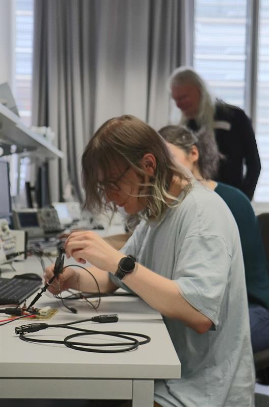 Eine Person arbeitet an einem technischen Arbeitsplatz in einem laborartigem Lernraum. Sie hantiert mit elektronischen Komponenten und Werkzeugen, darunter Prüfspitzen und Kabel, die mit einer Leiterplatte verbunden sind. Auf dem Tisch stehen Messgeräte wie Oszilloskope und Monitore. Im Hintergrund sind weitere Personen sichtbar, was auf eine kollaborative oder lehrende Umgebung hinweist. Die Szene zeigt praxisnahes Lernen oder Forschen im Bereich des Ingenieurwesens.