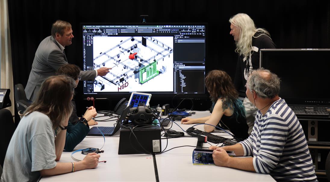 Sechs Personen sitzen oder stehen um einen Tisch in einem modernen Lernraum. Sie diskutieren ein technisches Raum-Modell, das auf einem großen Monitor angezeigt wird. Der Bildschirm zeigt ein 3D-Layout mit verschiedenen akustischen Bauelementen beschriftet mit „HSD“. Eine Person zeigt mit einem Zeigestab auf den Monitor und erklärt Details. Auf dem Tisch liegen technische Geräte wie ein VR-Headset, Laptops und Kabel. Die Szene vermittelt praxisnahes, kollaboratives Lernen in den Bereichen Technik, Robotik oder CAD.