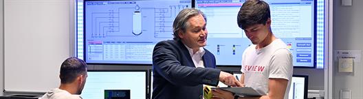 Ein Mann im Anzug zeigt einem jüngeren Kollegen Informationen auf einem Tablet in einem Kontrollraum mit großen Monitoren, die technische Systemdiagramme anzeigen.