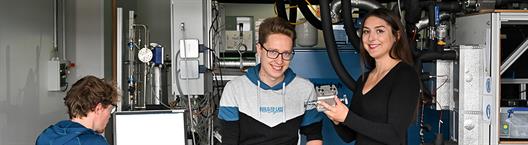 Eine Studentin und ein Student im Vordergrund des Bildes schauen in die Kamera. Sie befinden sich in einem Raum mit technischen Anlagen. Im Hintergrund sitzt ein Student vor einem PC und hat den Rücken zur Kamera gedreht.