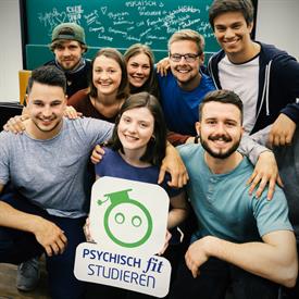 Young people group in front of a blackboard and hold up a sign with the title "Study mentally fit".