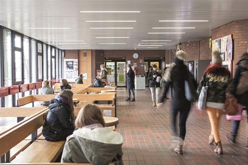 Studierende auf einem Flur der HSD. Links sitzend zwei Studierende auf Bänken und rechts im Bild gehen drei Studentinnen vorbei.