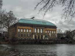 Das Foto zeigt das Museum K21 Ständehaus in Düsseldorf. Das historische Backsteingebäude liegt malerisch am Kaiserteich und ist durch seine große, moderne Glaskuppel markant geprägt. Vor dem Eingang ist eine Reiterstatue zu sehen, während sich im Vordergrund das Wasser des Teiches spiegelt. Die winterlich kahlen Bäume und der graue Himmel verleihen der Szene eine eher ruhige, etwas melancholische Stimmung.