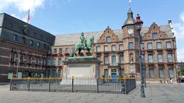 Das Foto zeigt das Düsseldorfer Rathaus am Marktplatz. Im Vordergrund steht das Reiterdenkmal von Jan Wellem, umgeben von einem schmiedeeisernen Zaun. Dahinter erhebt sich das historische Backsteingebäude mit Uhrturm, vielen Fenstern und blumengeschmückten Fassaden.