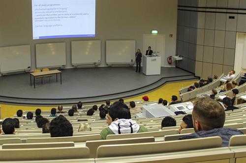 In a big auditorium. The view from high up down to the podium, where 2 women are giving a presentation. The seats of the auditorium are filled with refugees, who are interested to study.