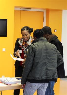 An dark skinned staff member of HSD, dressed in a black jacket and a colourful scarf, is advising 2 interested refugees about studying at HSD. They are standing in front of the information stand, which is set up in front of a yellow wall. The woman is pointing at a brochure.