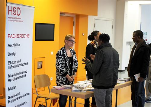 Infostand der HSD mit Roll-Up und Broschüren ist vor einer gelben Wand aufgebaut. 2 Mitarbeiterinnen des International Office beraten 2 dunkelhäutige Flüchtlinge.