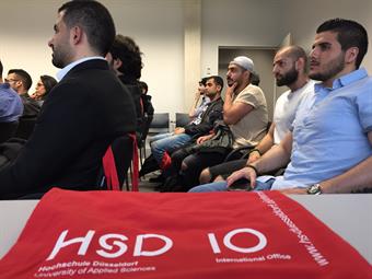 Junge Männer sitzen auf Stühlen in Reihen.
Im Vordergrund liegt einen rote Jute-Tasche mit der Aufschrift HSD IO auf einem Tisch.