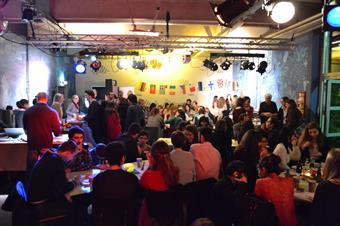A big room, which is colourfully light and decorated with different national flags. There are many tables filled with international students. They are eating and drinking.