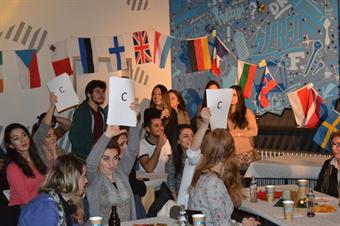 Many international students are sitting at tables. They are holding answer sheets up for a quiz.