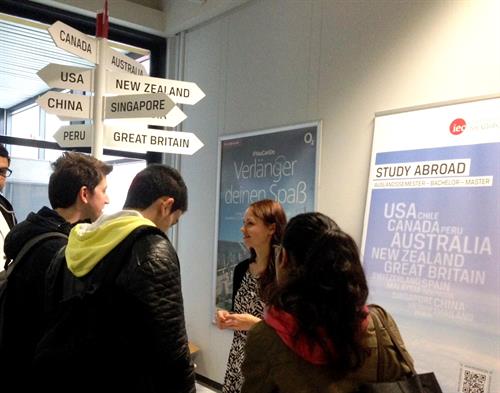 3 students are standing in front of a desk. Behind the desk a young women, dressend in a black and white dress - gives advice. The the background we see a blue roll-up with names of different countries on in and a guide post, which shows the direction of different countries.