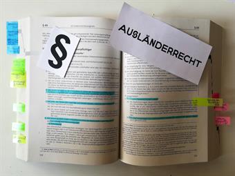 Ein dickes Buch mit Gesetztestexten ist aufgeschlagen. Manche Sätze sind markiert, viele Seiten sind mit Fähnchen markiert. Es liegt ein großes Paragraphen-Zeichen und das Wort Ausländerrecht auf dem Buch.