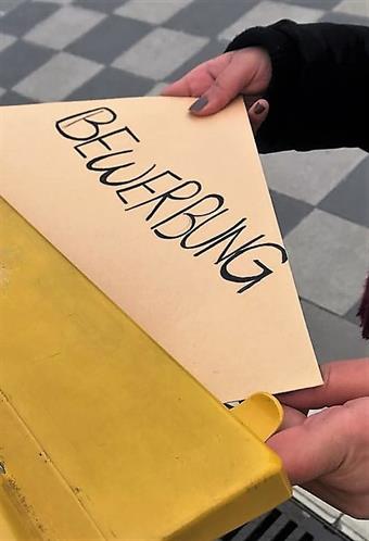 Someone is putting a big envelope with the word "Bewerbung" written on it, into a yellow Mailbox.